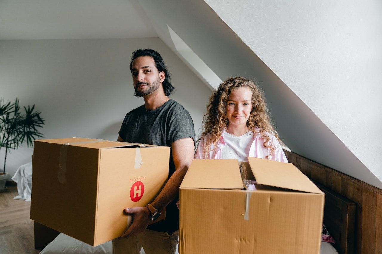 Couple carrying moving boxes in an attic, smiling, ready to relocate.