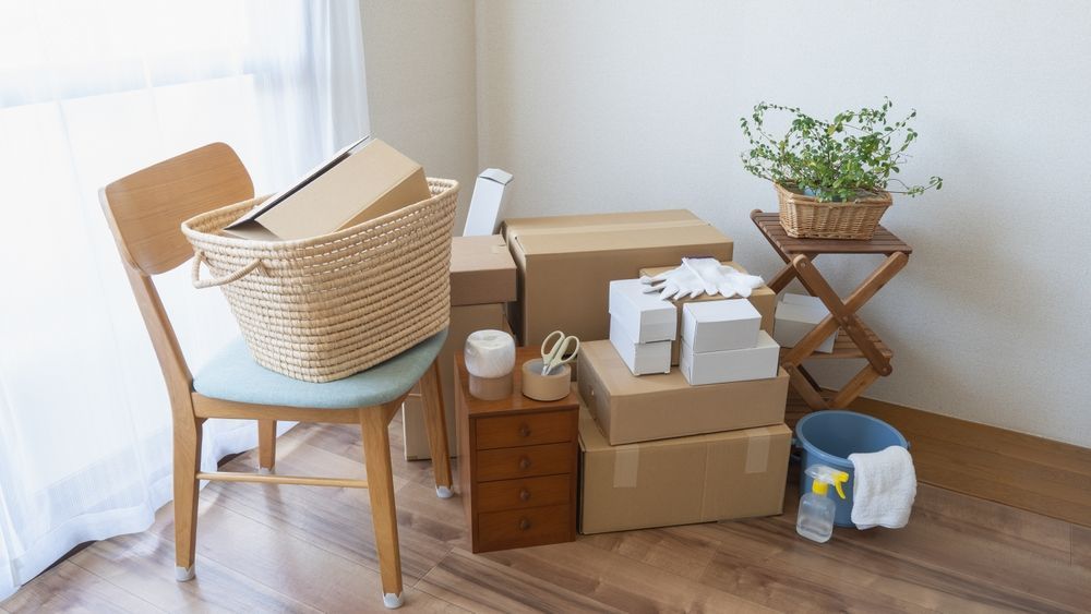 Packed moving boxes and household items in a bright corner by a chair and plant stand.