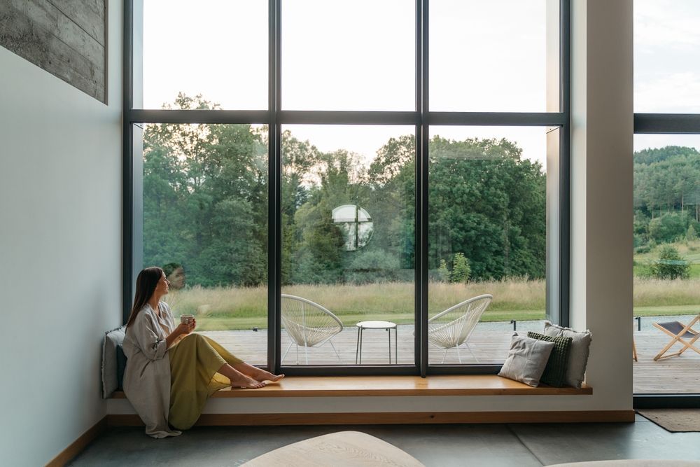 Person sitting on a window seat by large floor-to-ceiling windows overlooking a green garden