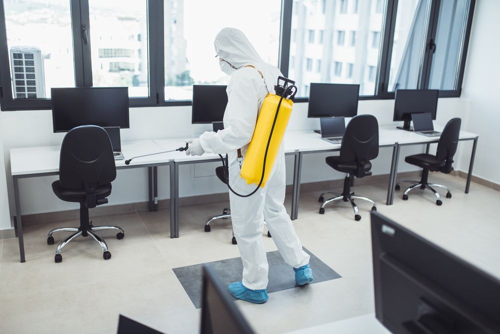 Worker in white hazmat suit disinfects a modern office with a yellow backpack sprayer.