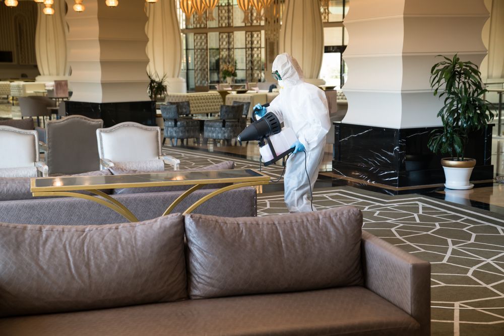 Person in protective suit disinfecting a hotel lobby with chairs, sofas, and patterned carpet