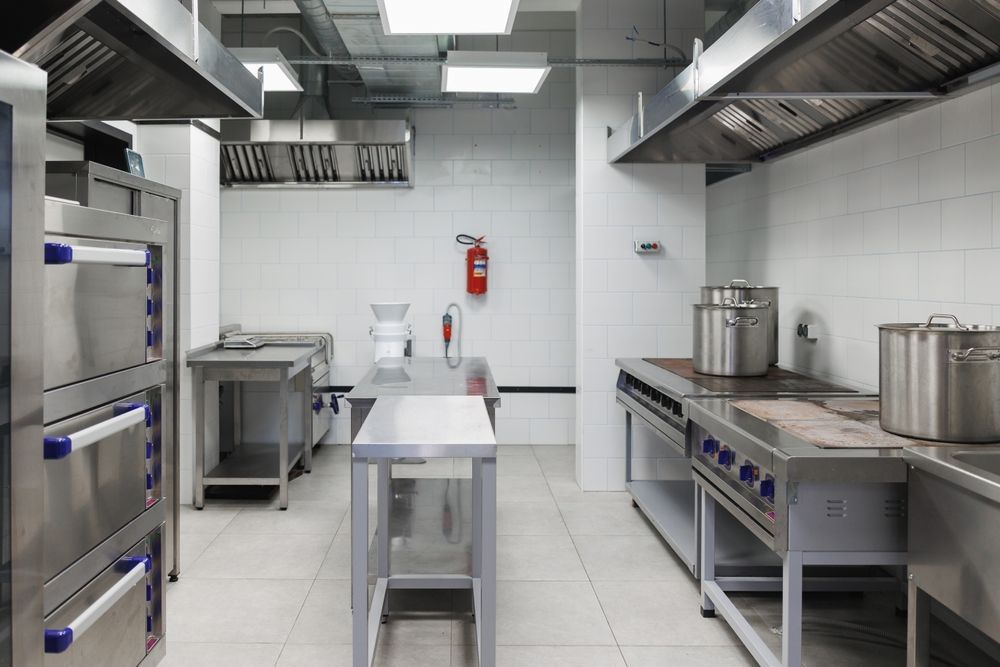 Stainless steel commercial kitchen with counters, ovens, pots, and a fire extinguisher on tiled walls.