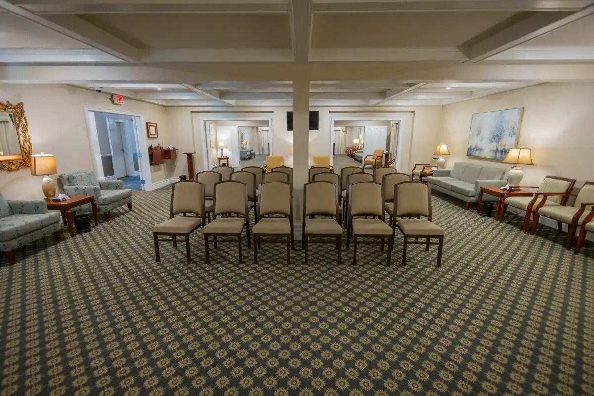Interior of a funeral home chapel with rows of chairs, a patterned carpet, and soft lighting.