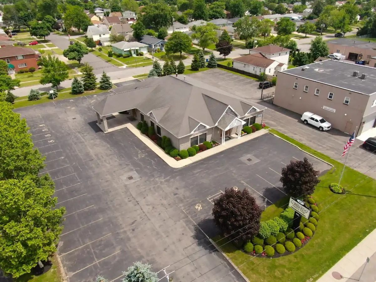 Aerial view of a gray building with a large parking lot, surrounded by green trees and houses.