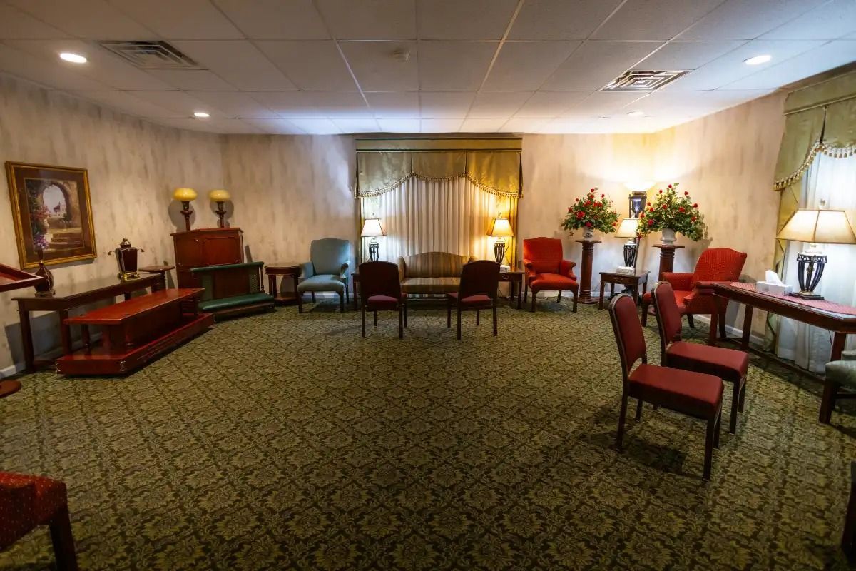 Funeral home interior with chairs, floral arrangements, and casket on green patterned carpet.