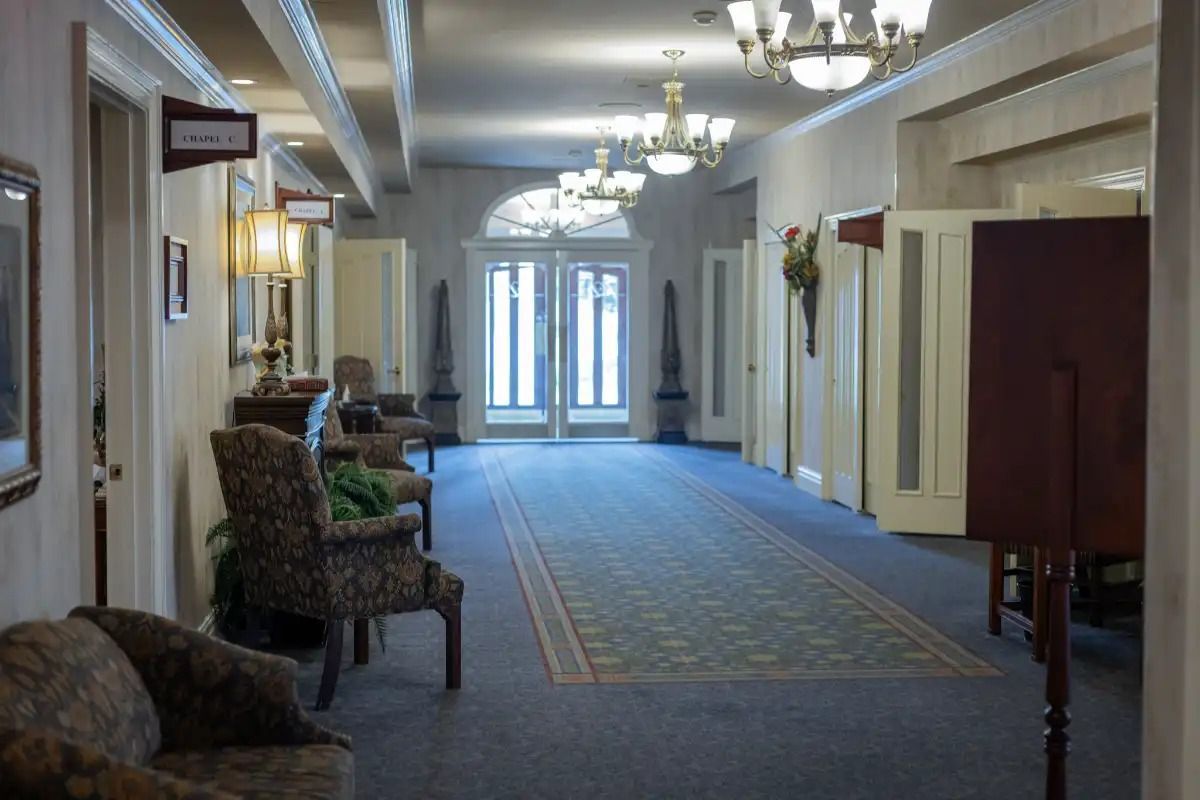 Long hallway with blue carpet, chairs, doors, and chandeliers. Double doors at the end with sunlight.