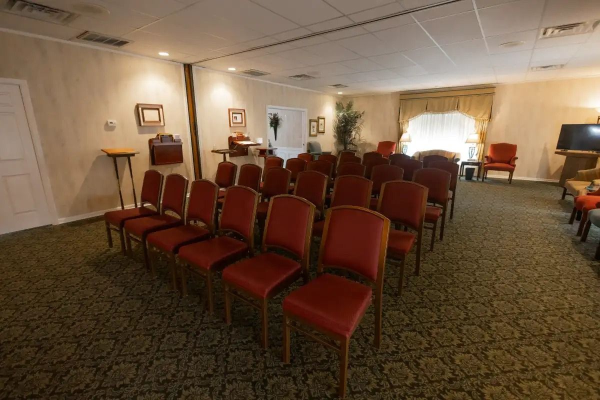 Rows of red chairs face a podium in a room with tan walls and floral carpet, likely a chapel.