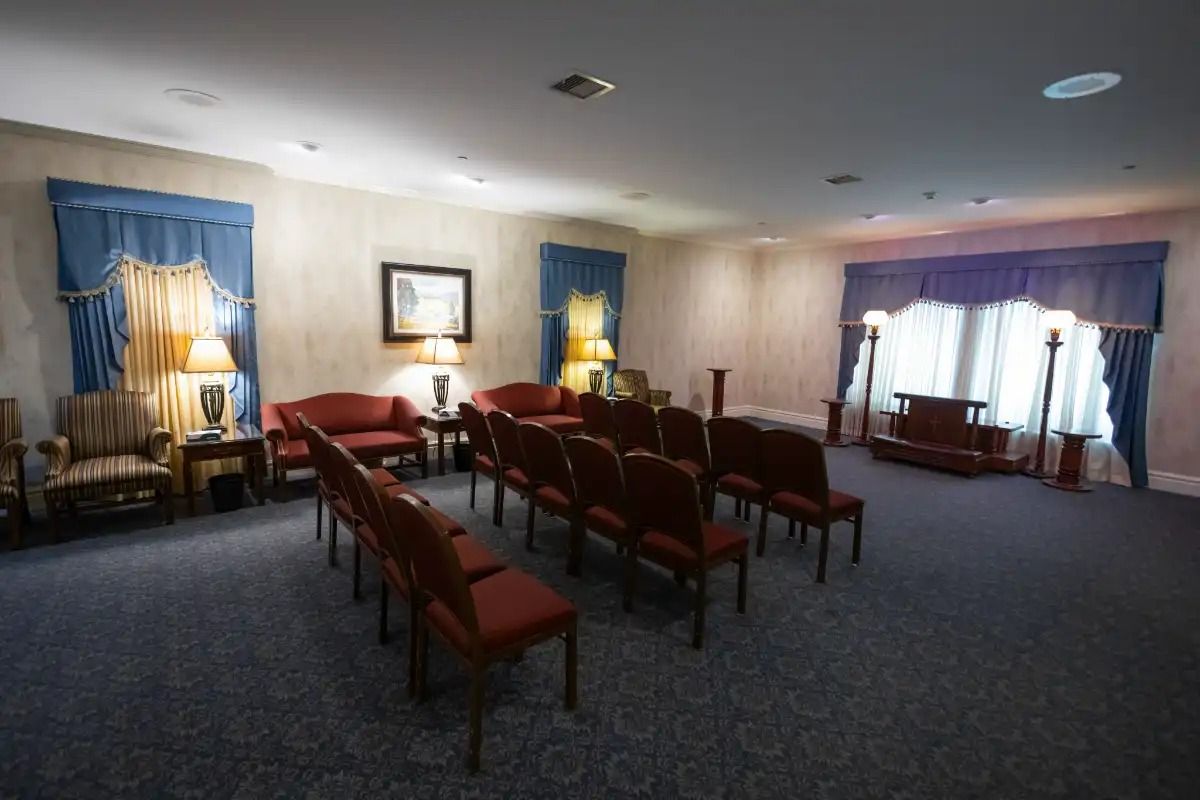 Interior of a funeral home chapel with rows of red chairs, blue curtains, and soft lighting.