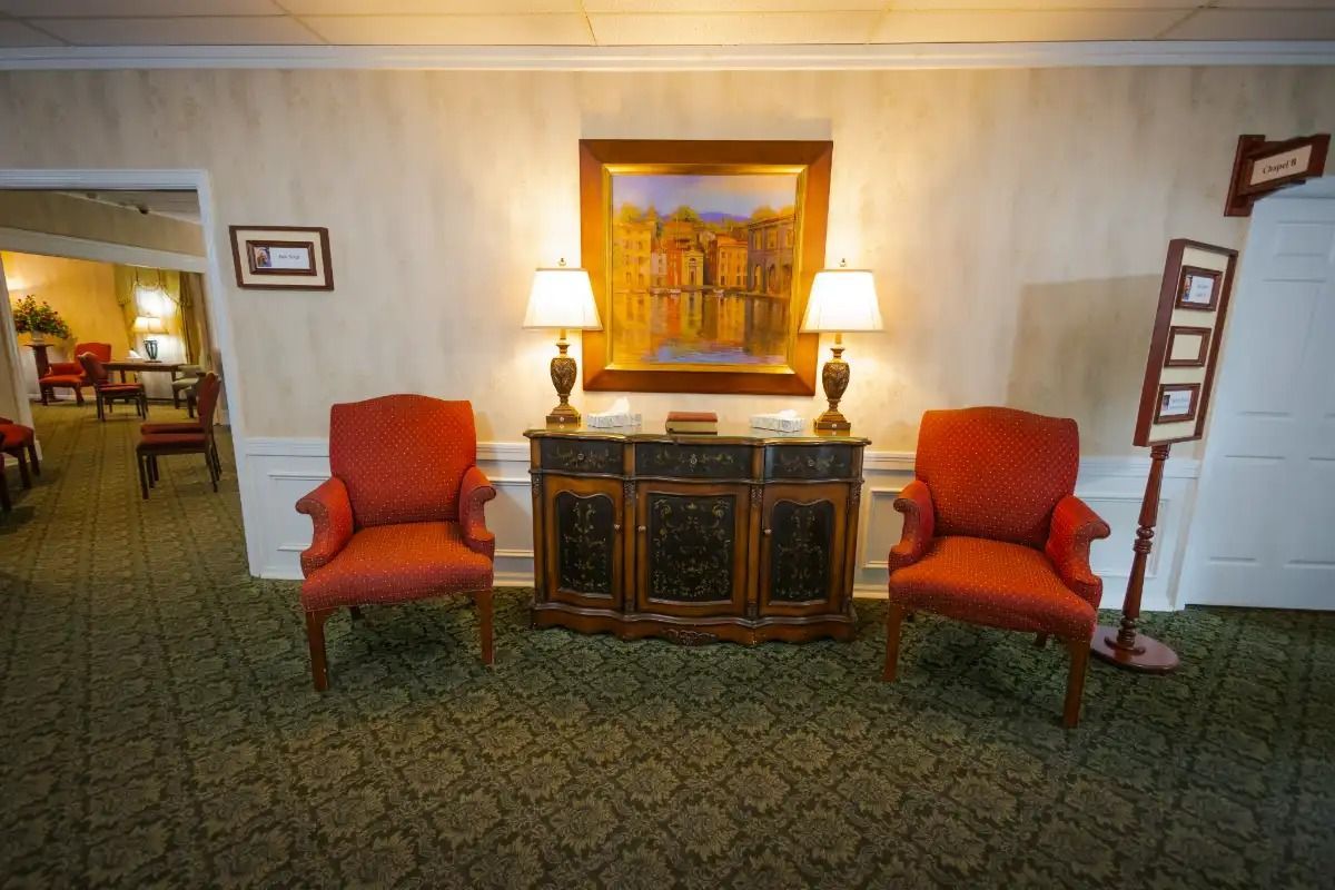 Two red armchairs flanking a dark cabinet, under a painting, with two lamps.
