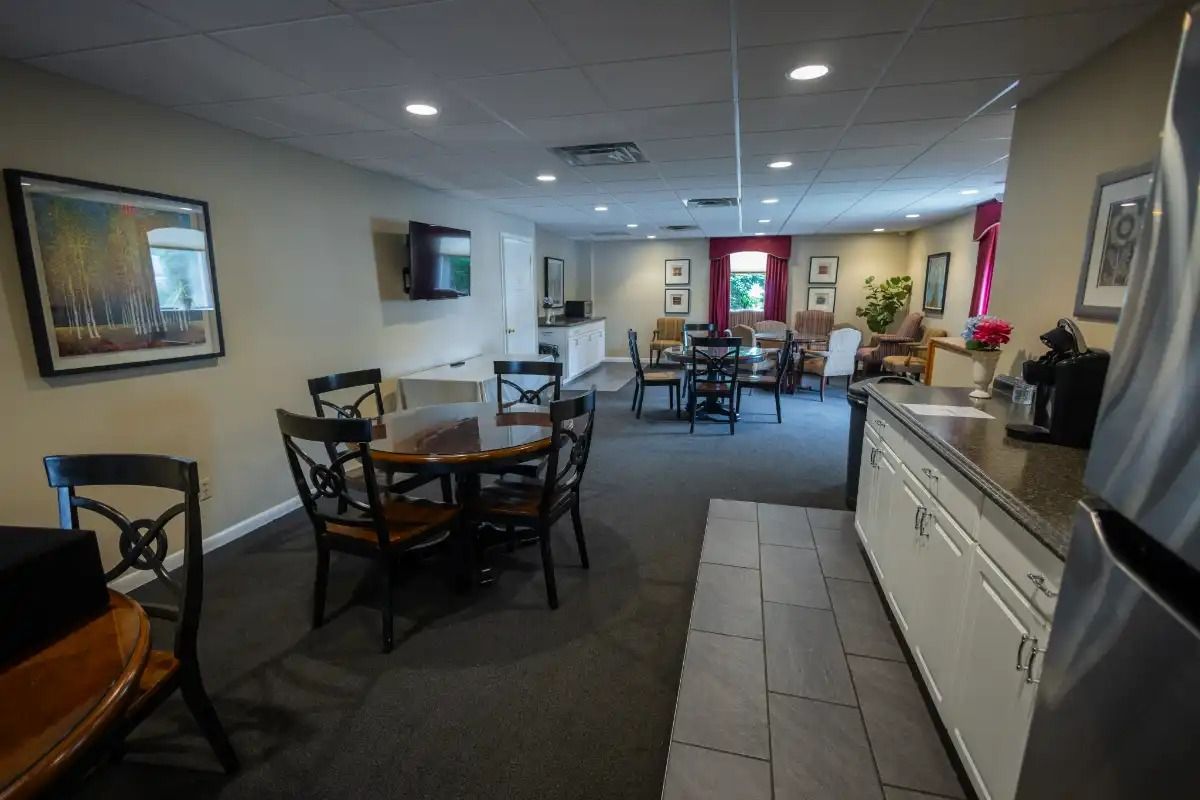 Common area with tables, chairs, and a kitchenette. Neutral walls, dark carpet, and a small TV.
