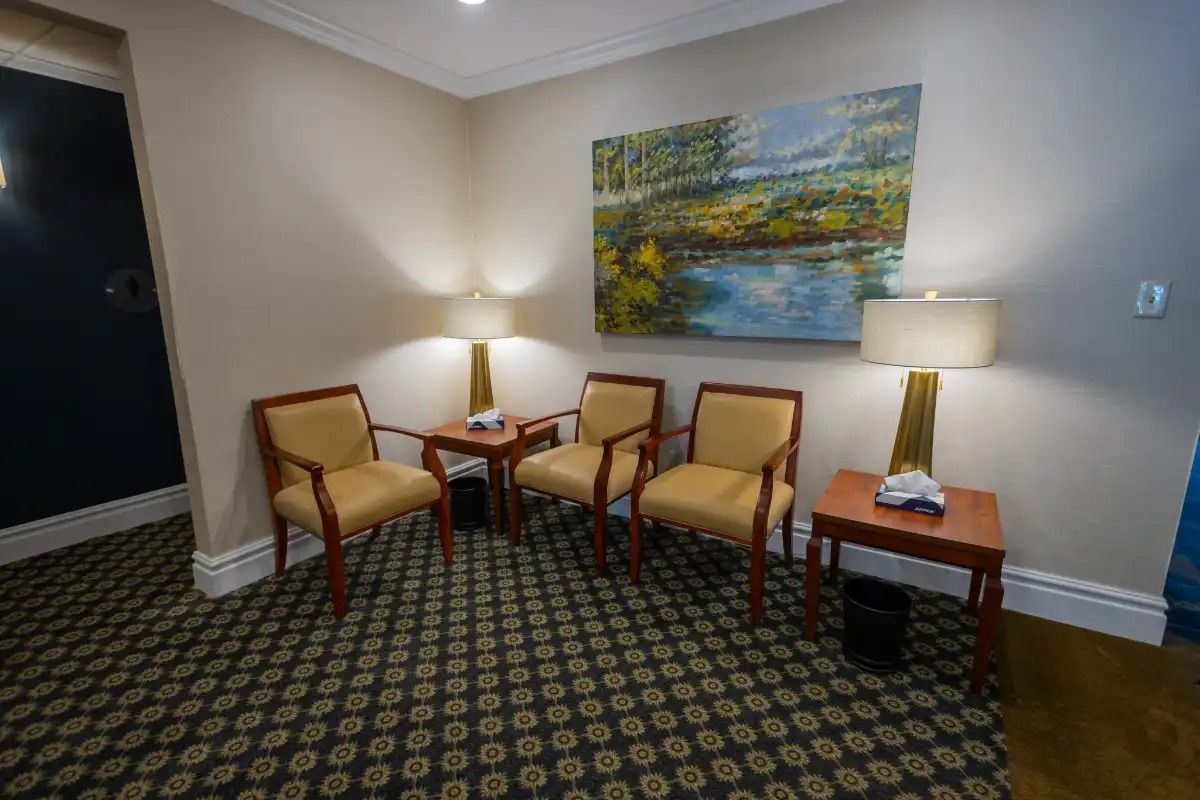 A waiting area with four chairs, two side tables, and a landscape painting. Beige walls and patterned carpet.