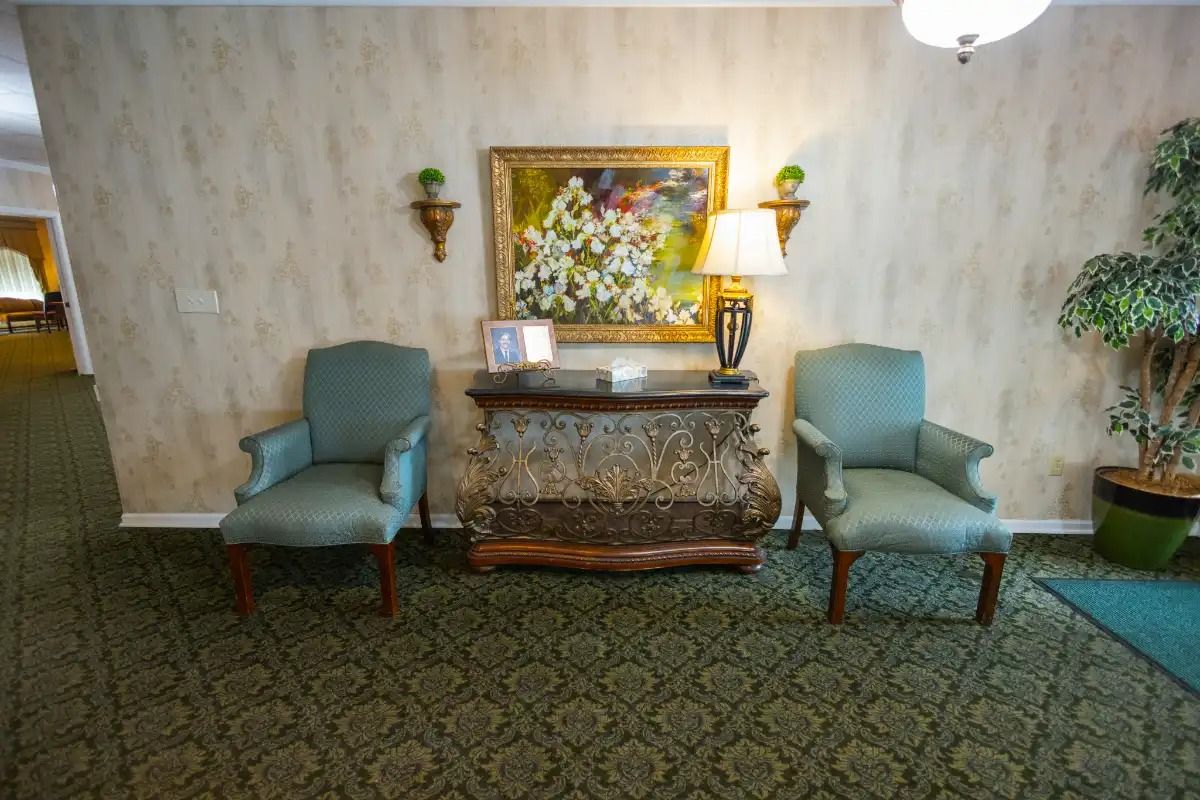 Two teal armchairs flank a decorative wooden console, topped with a lamp and picture, beneath a floral painting on a patterned carpet.