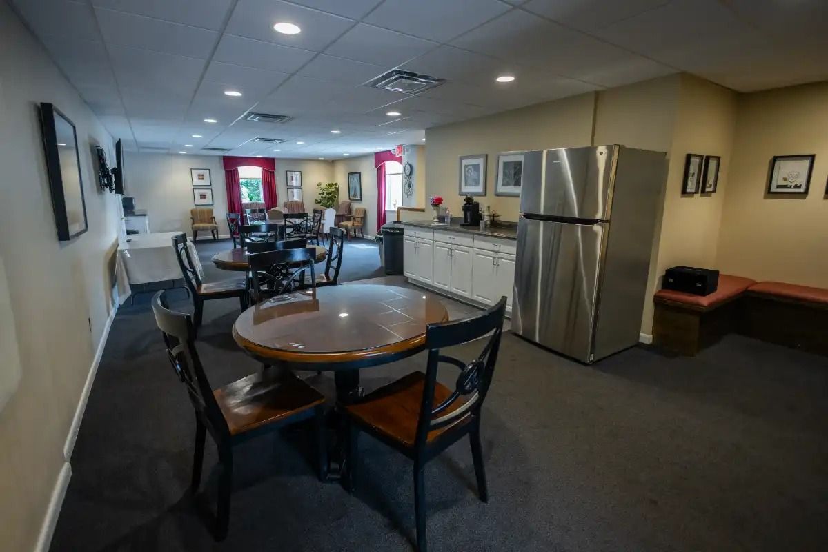 Interior of a room with round tables, chairs, a refrigerator, and a small kitchen area.