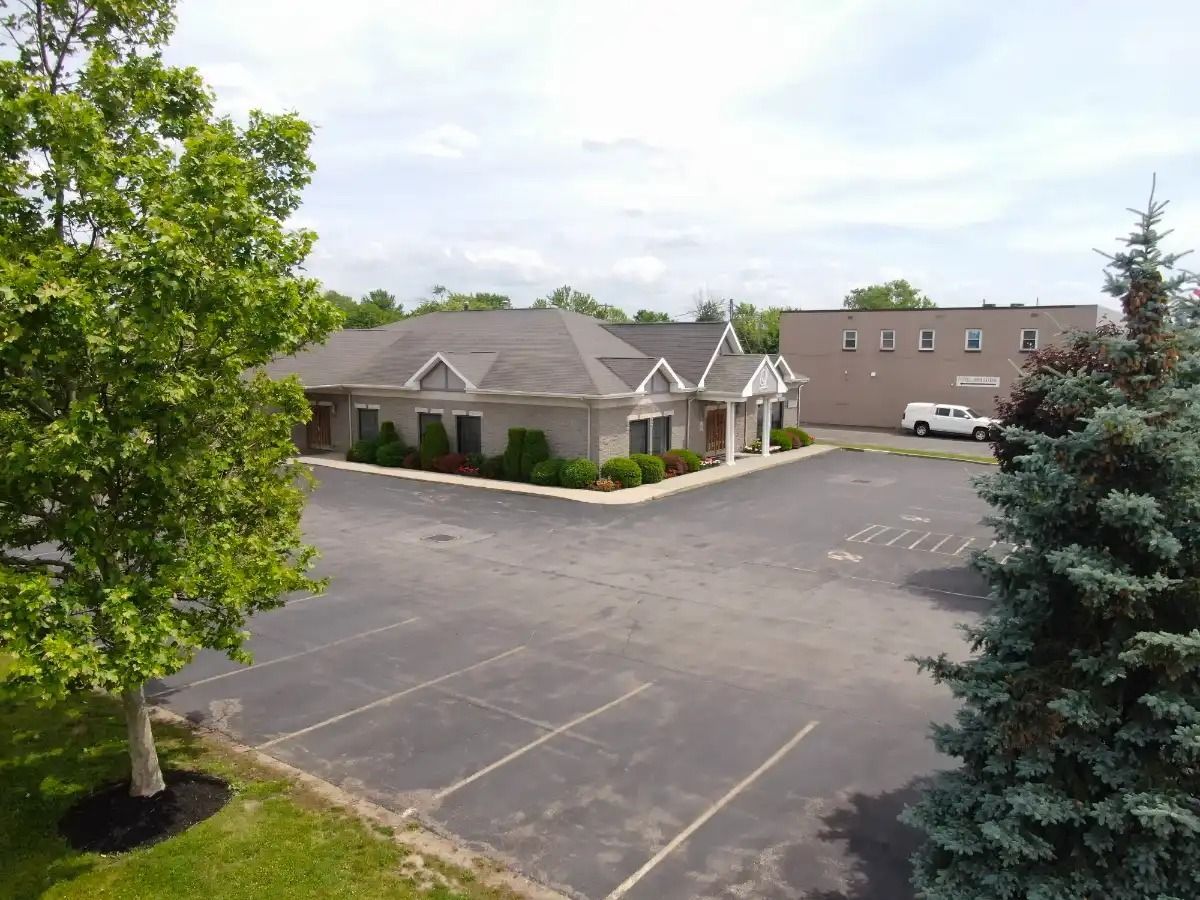 An aerial view of a gray brick building with a parking lot and trees on a sunny day.