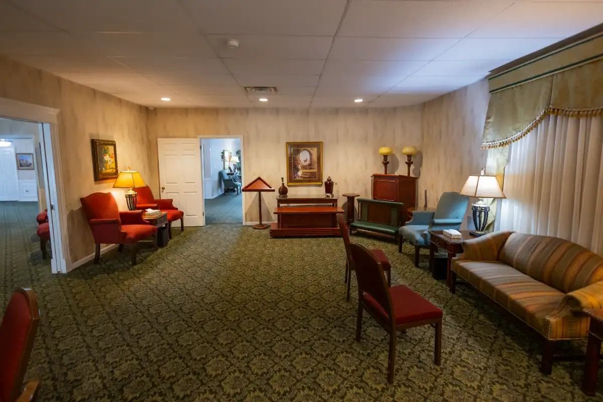 A funeral home's sitting room with chairs, sofas, and a piano. Carpet is green with a floral pattern.