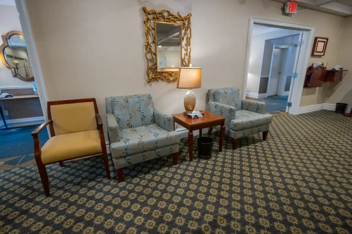 Lobby with chairs: yellow and patterned blue, small table with lamp, ornate mirror, carpet.