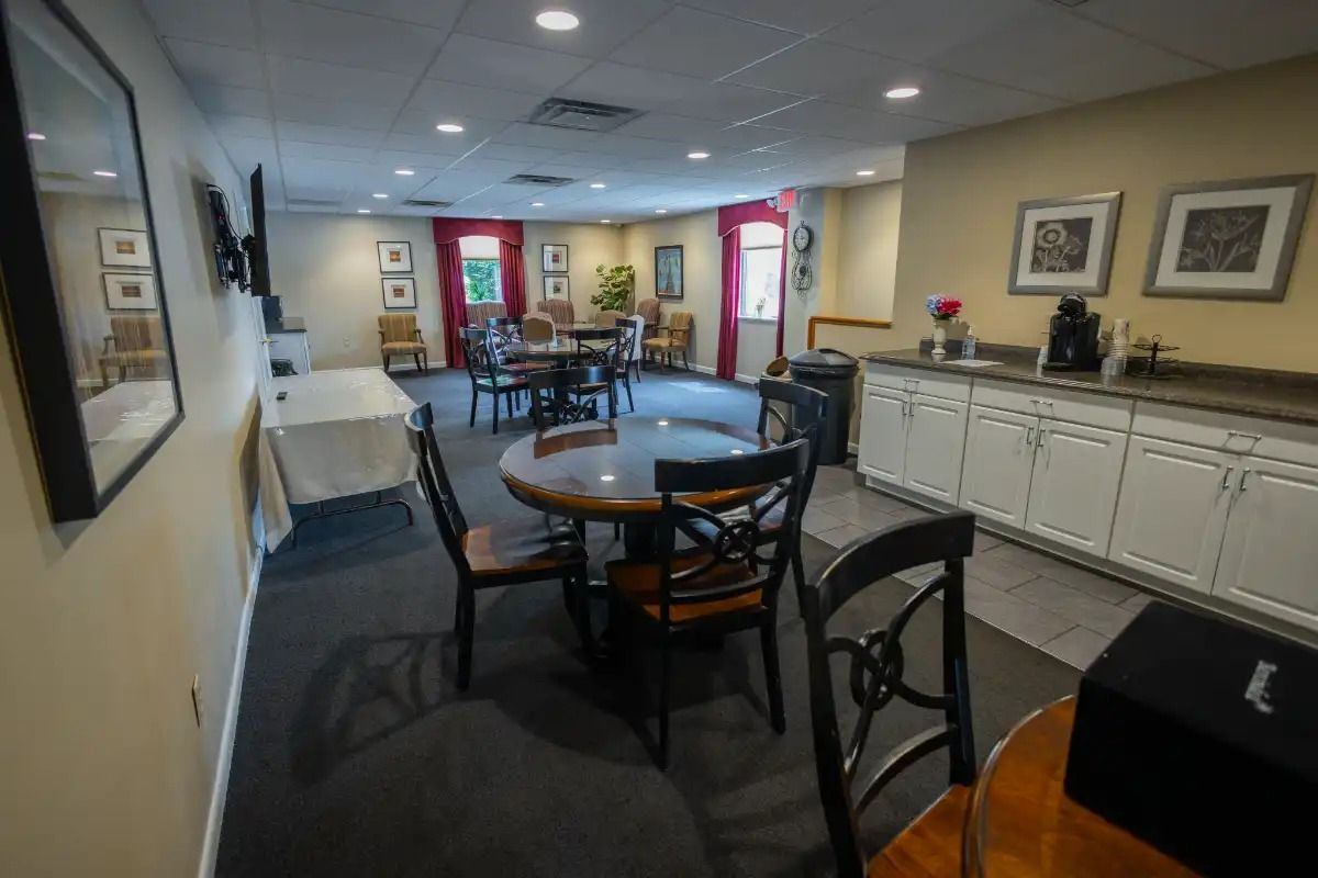 A room with round tables, chairs, and a coffee station; tan walls, dark carpet, framed art.