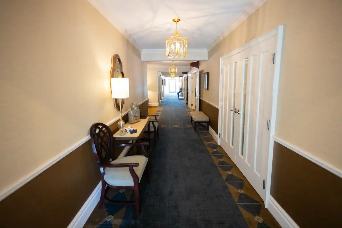 Long hallway with cream and brown walls, ornate lighting, and a table and chair.