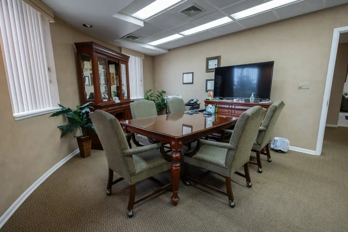 Meeting room with table, chairs, cabinet, TV, and window. Beige walls, carpet; neutral tones.