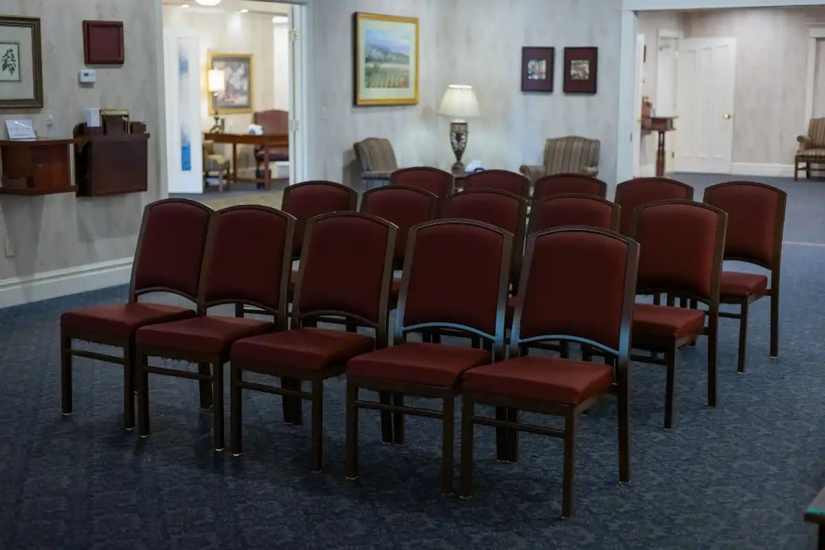 Rows of empty burgundy chairs in a room, presumably for a gathering, with artwork and a doorway visible.