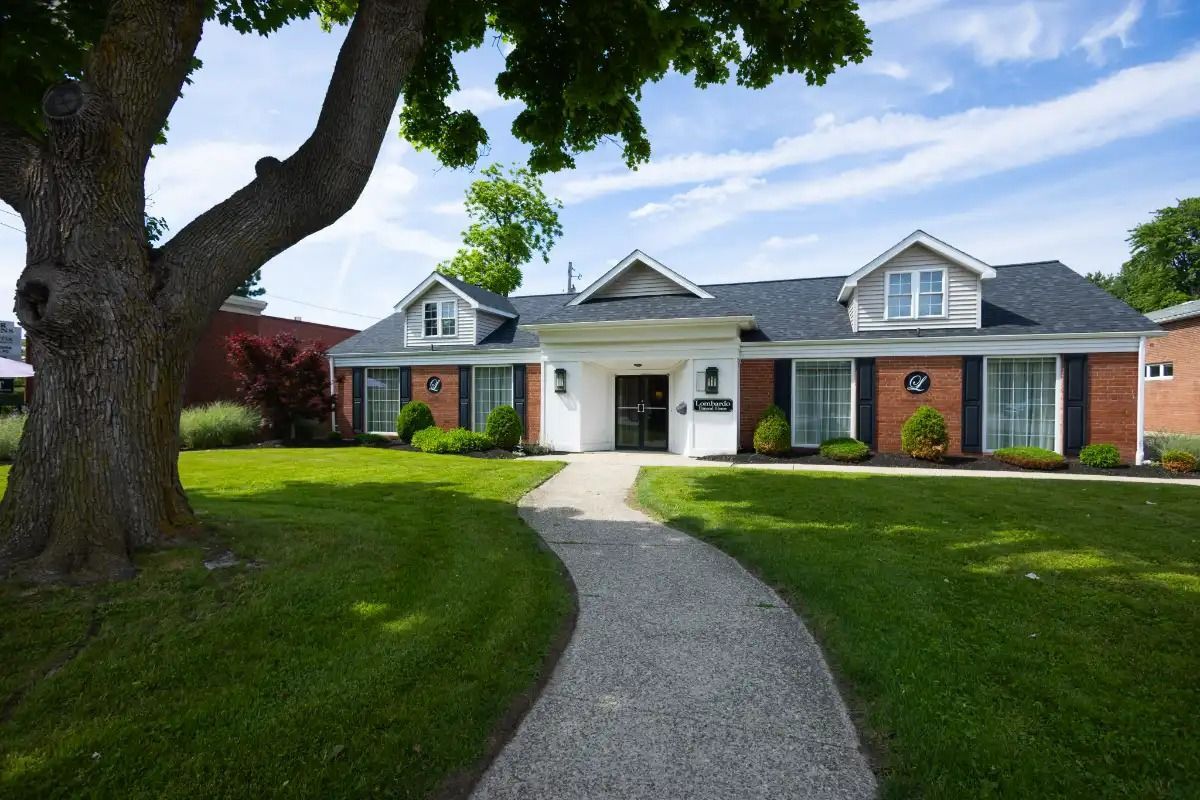 White building with black shutters, dormers, and a curved path through green grass.
