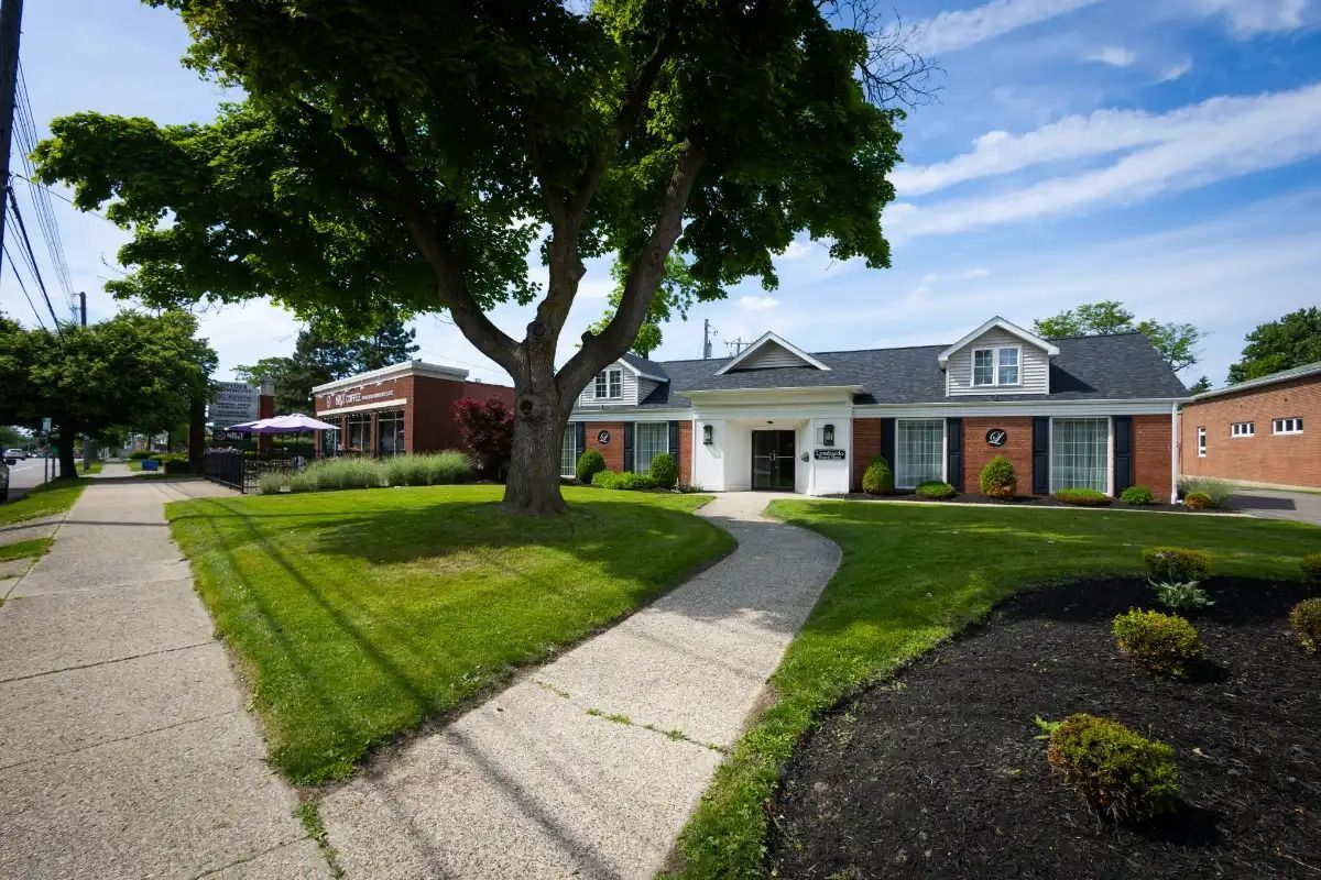 A white building with black shutters and a dark roof, on a green lawn with a sidewalk, next to a street.