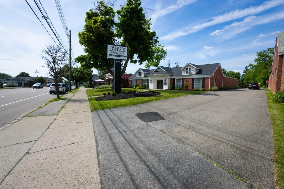 Street view of a building with a sign in front, asphalt parking lot, and sidewalk on the side of a road.