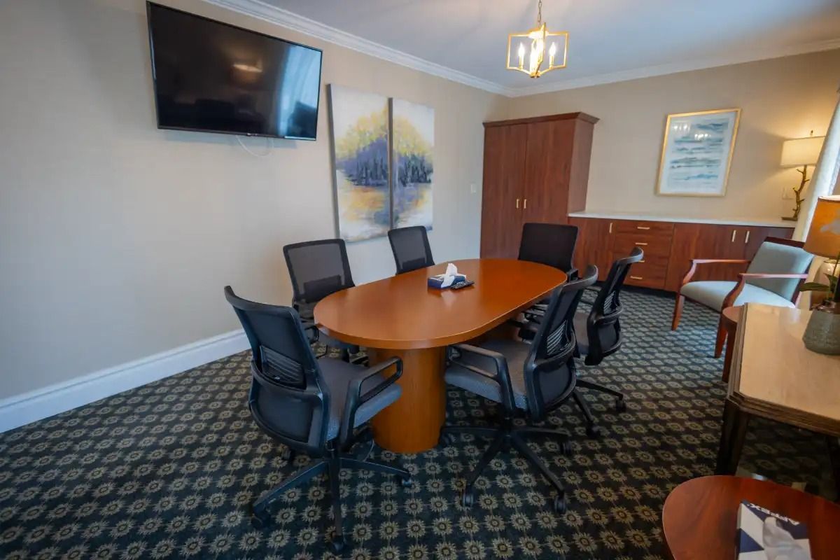A conference room with an oval table, chairs, artwork, a TV, and cabinet. Beige walls and patterned carpet.
