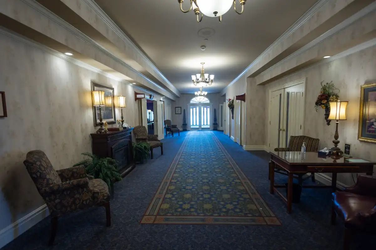 Long hallway with blue carpet, chairs, lamps, and art on walls leading to a bright doorway.