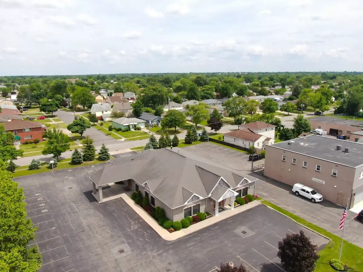 Aerial view of a gray building with a long roof, surrounded by a parking lot, trees, and houses on a sunny day.