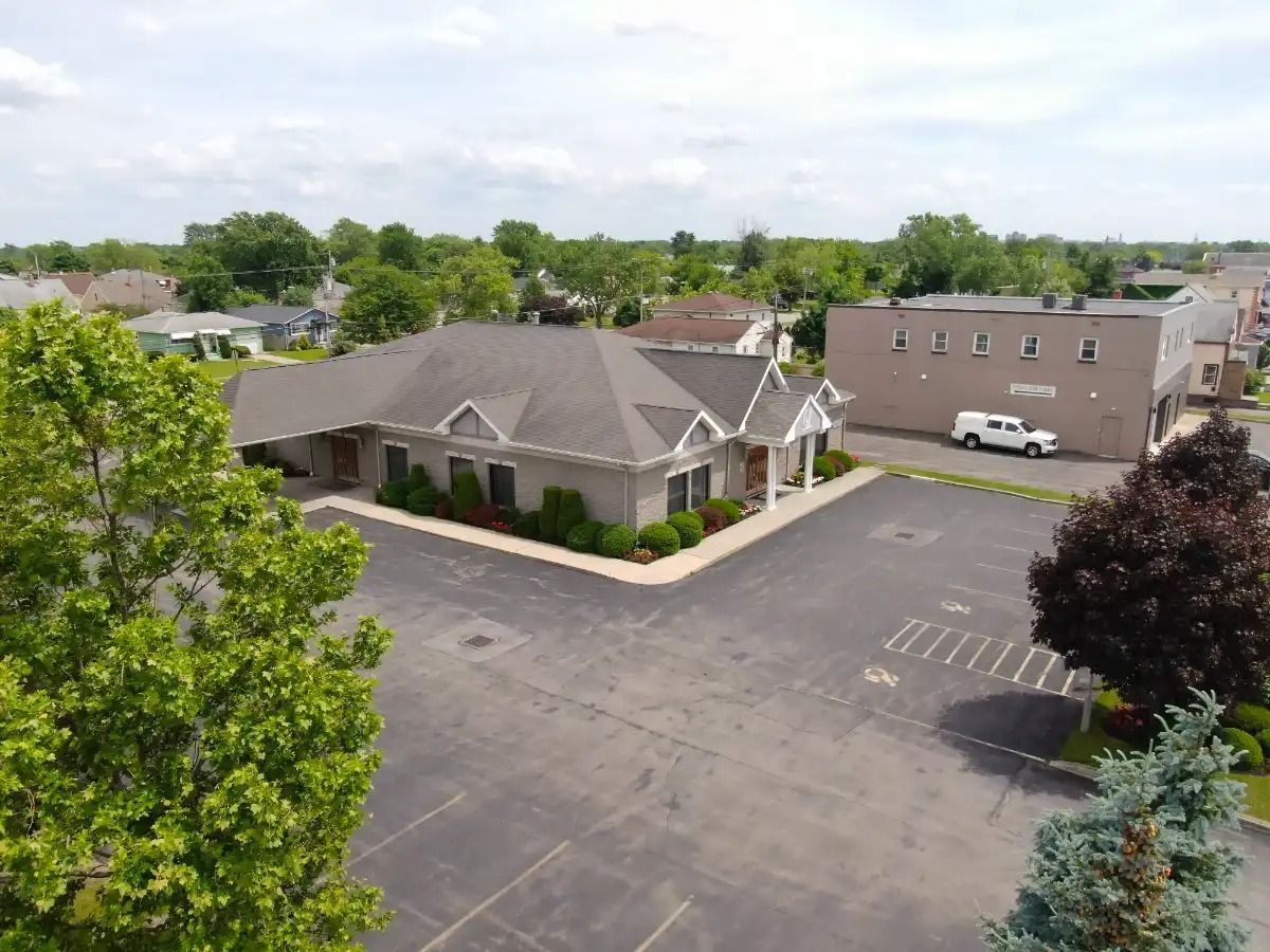 Aerial view of a beige building with a gray roof and parking lot, trees in the foreground, and other buildings in the background.