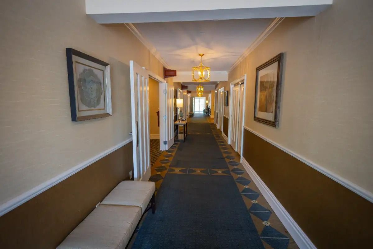 Long hallway in a building, with doors, art, a bench, and a blue carpet.