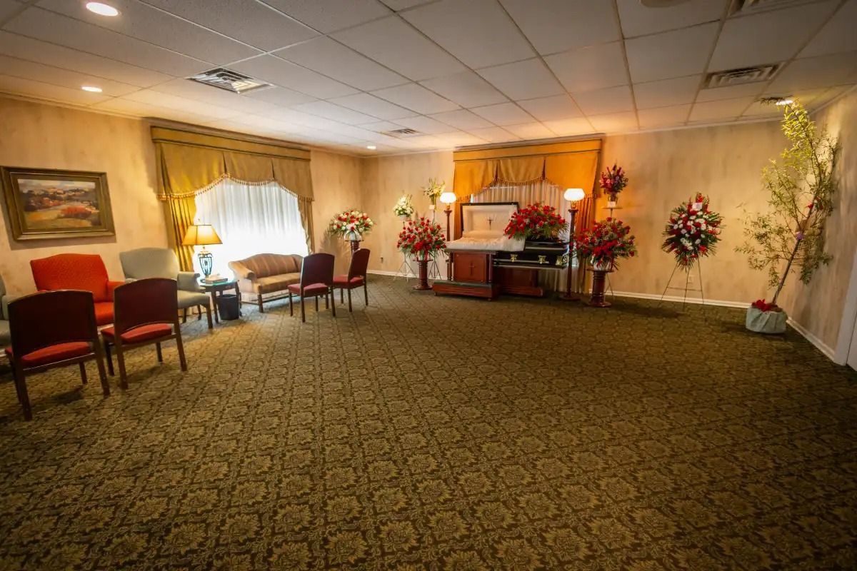 Funeral home interior with chairs, floral arrangements, and a casket area.