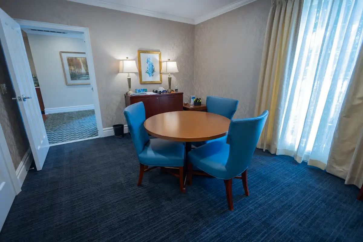A round table with four blue chairs in a room with blue carpet and a window with curtains.