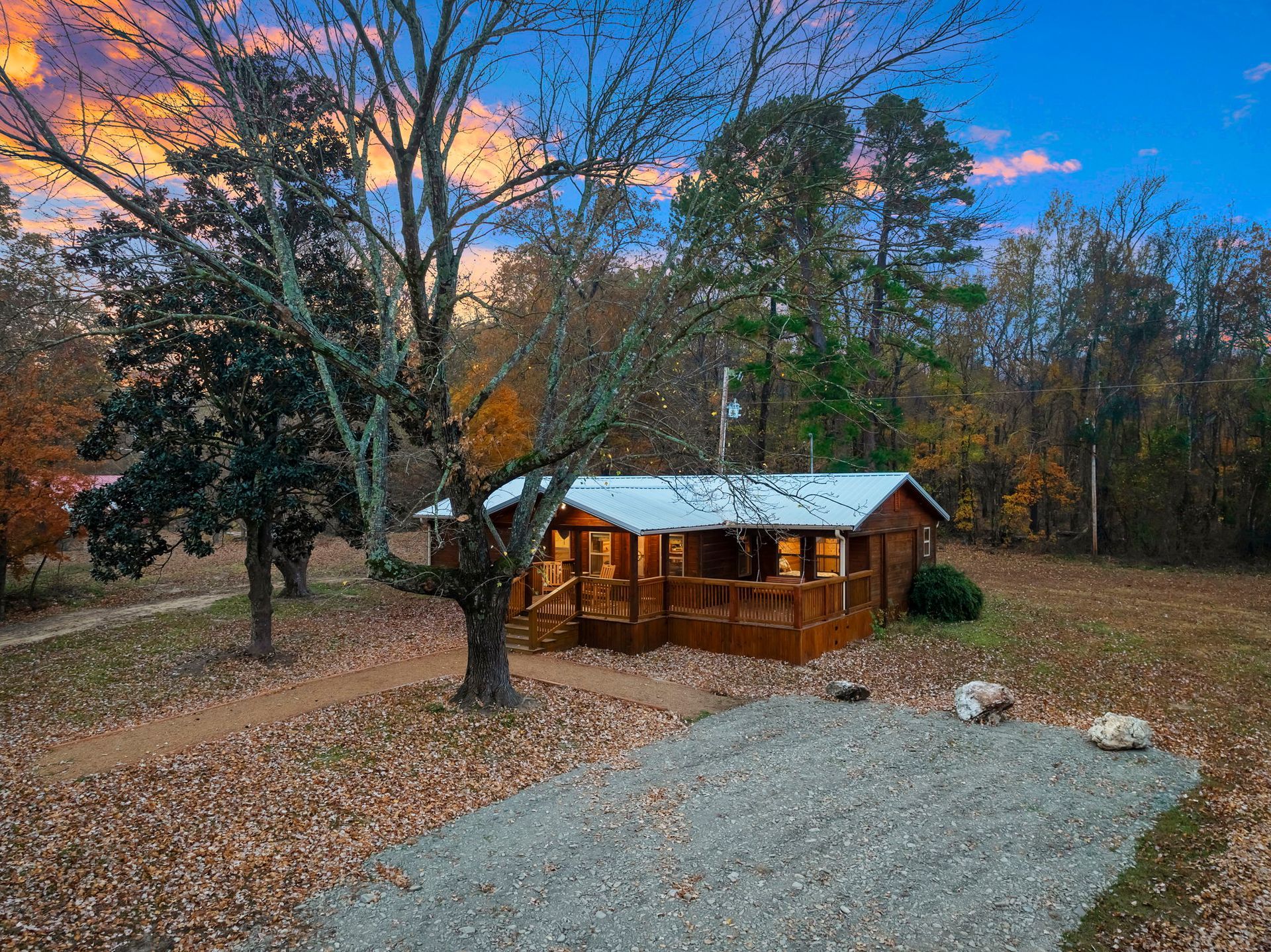 A small wooden cabin with a metal roof sits among trees during sunset, with a gravel area in the foreground.