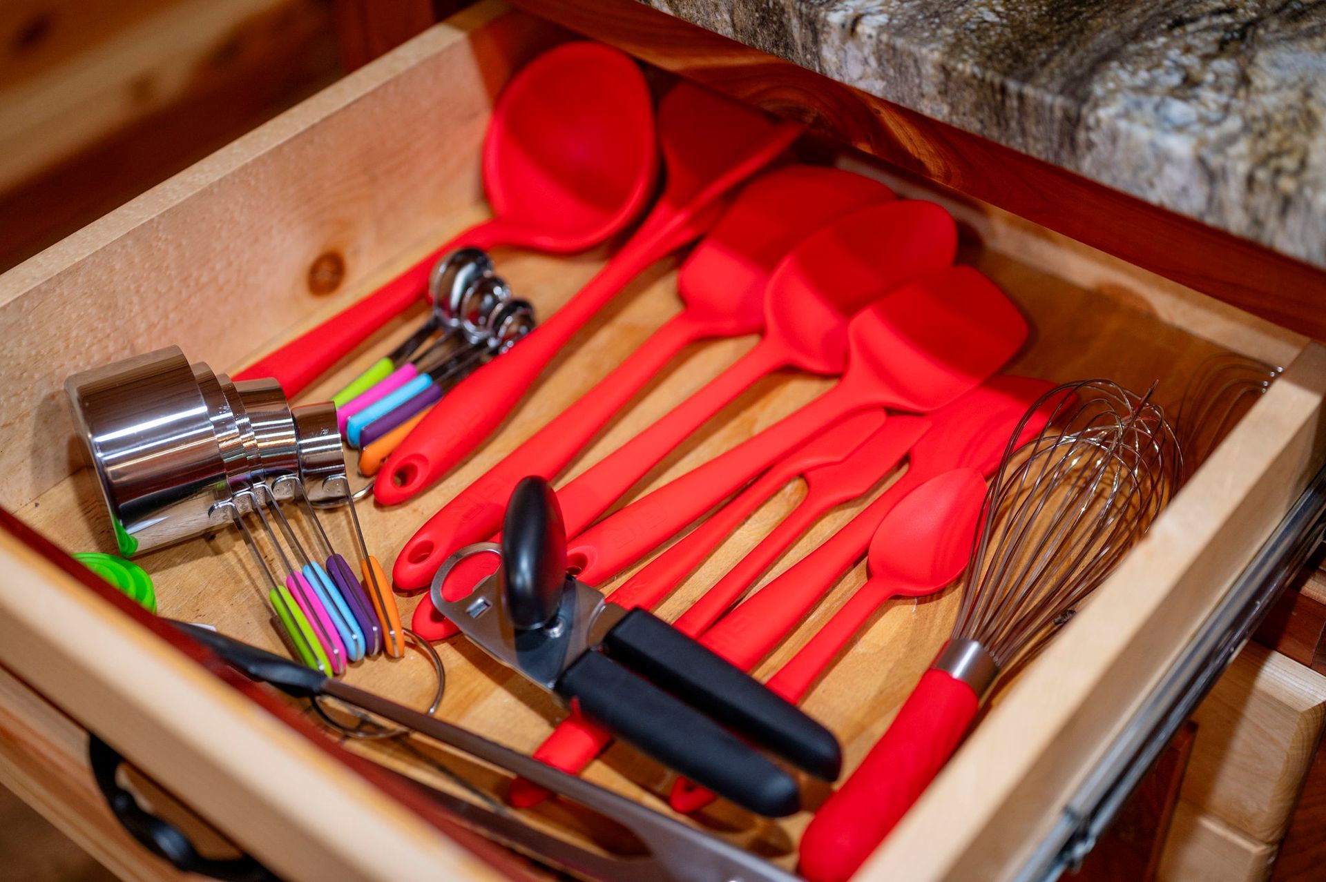 A wooden kitchen drawer filled with an assortment of red silicone cooking utensils, a whisk, a can opener, and tools.