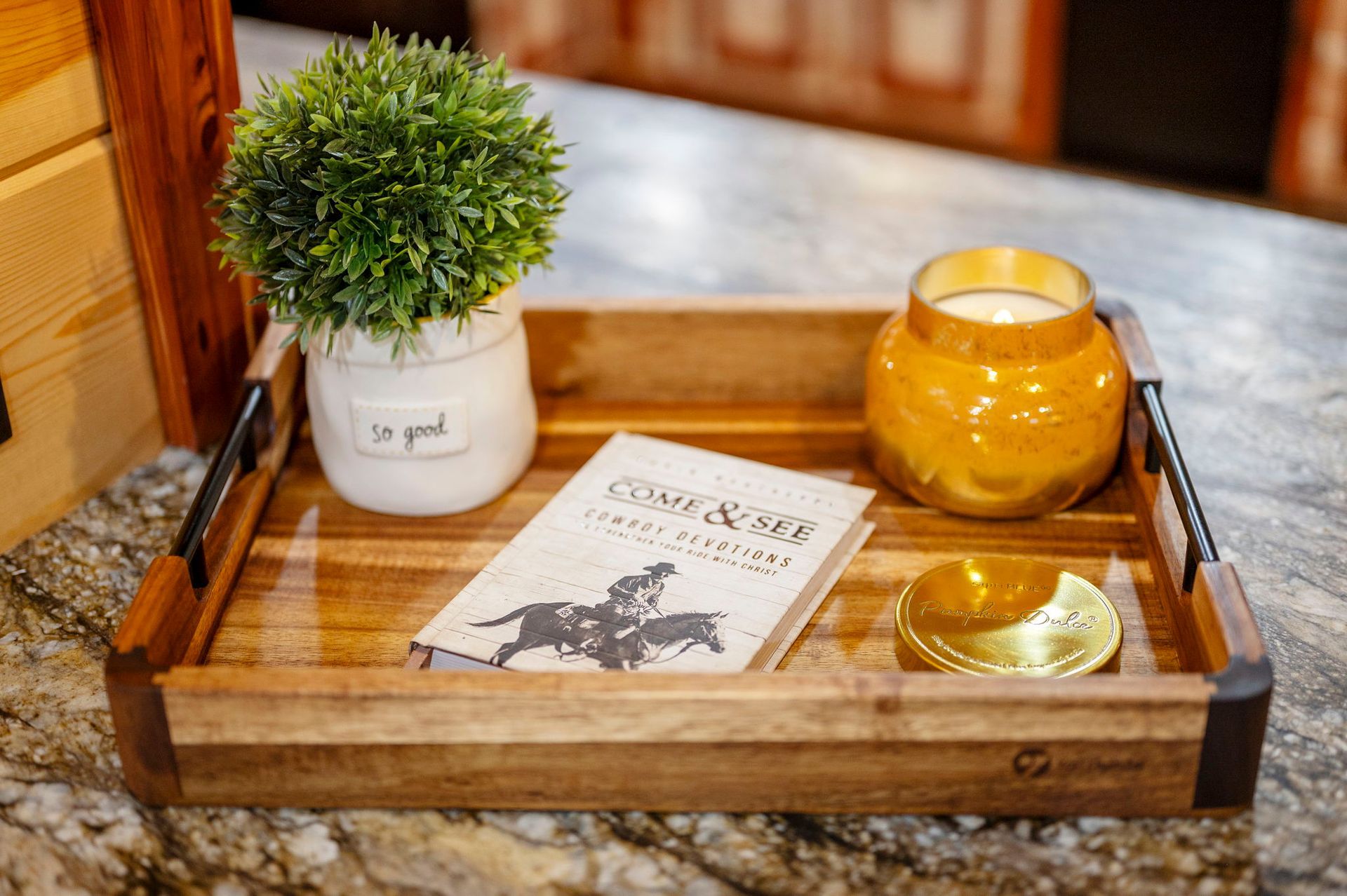 A wooden serving tray holds a small potted plant, a candle in a mustard-yellow jar, and a book on a granite surface.