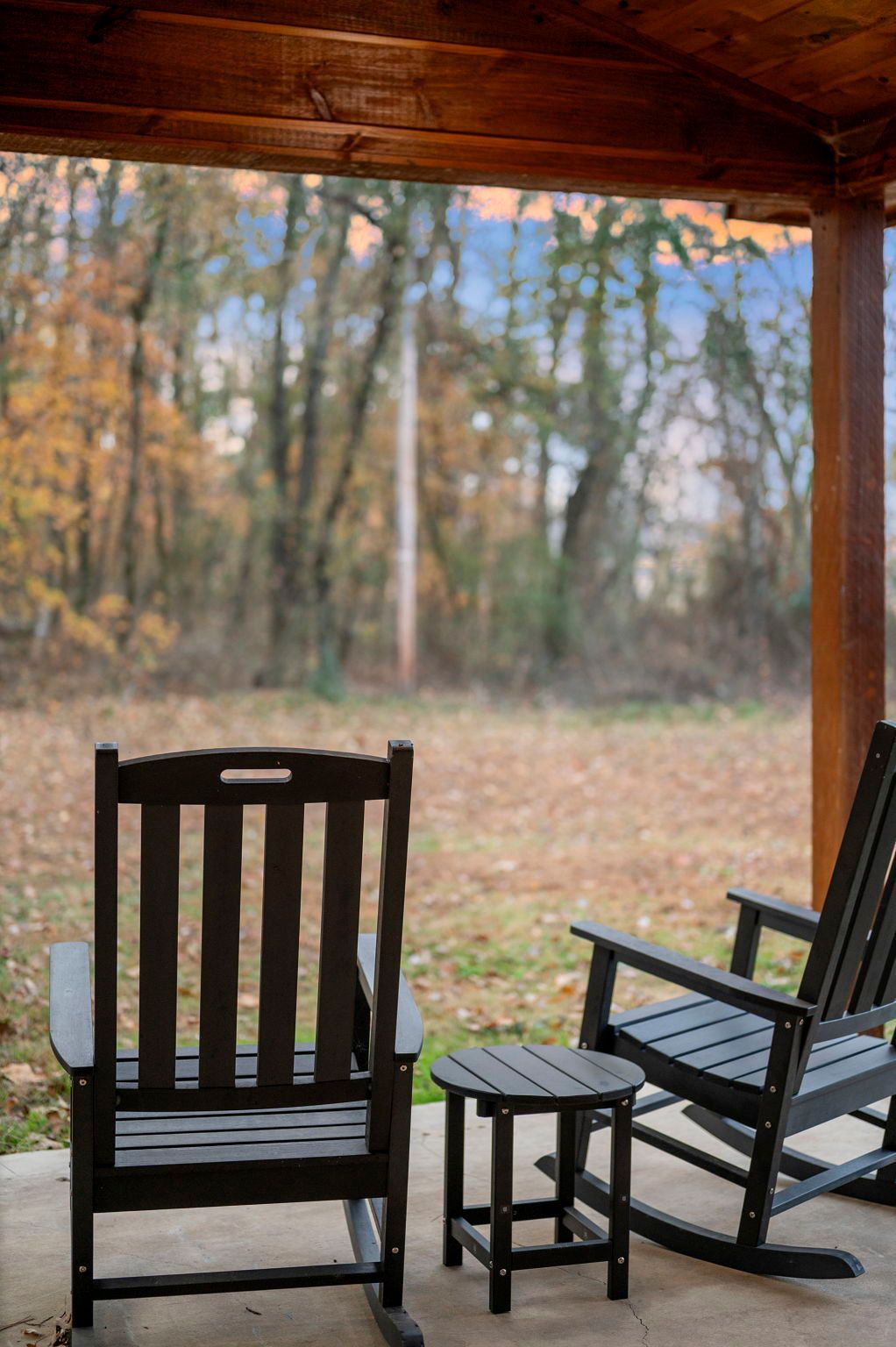 Two black rocking chairs and a small matching side table on a wooden porch overlooking a wooded area in autumn.