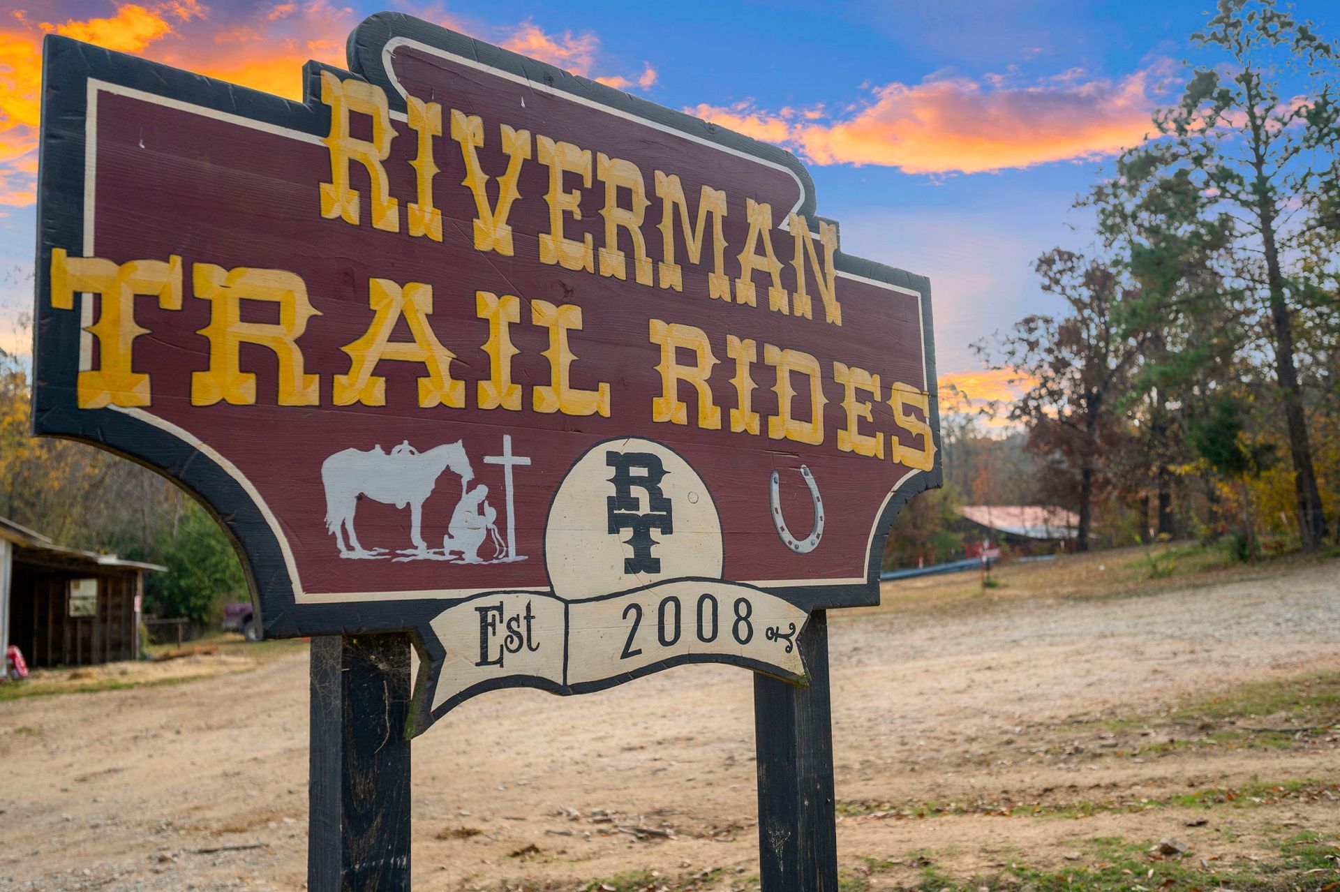 A wooden sign for Riverman Trail Rides, est. 2008, featuring a silhouette of a person kneeling by a horse against a sunset.