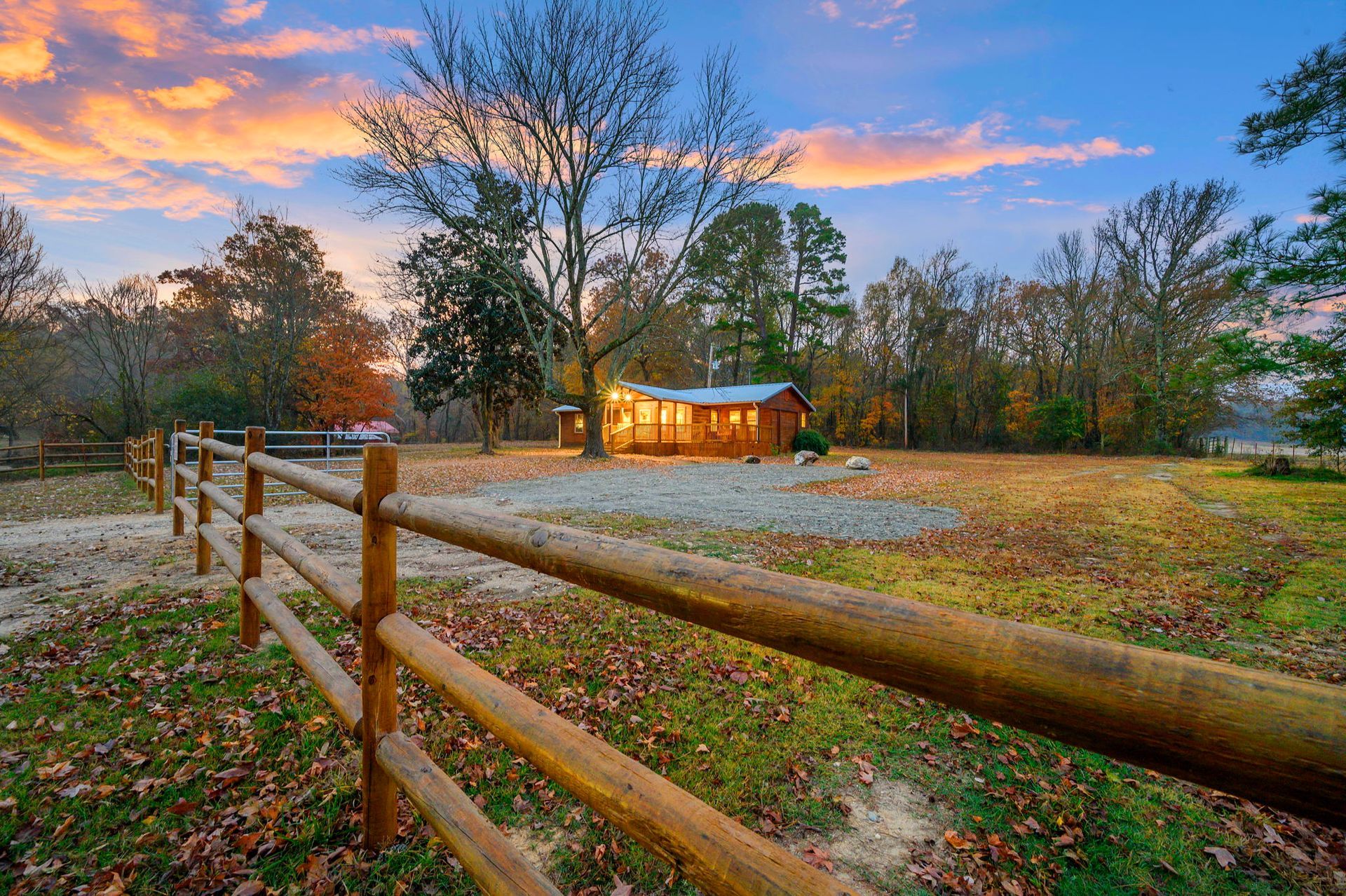 A wooden cabin glows warmly at twilight, viewed across a grassy field bordered by a rustic wooden split-rail fence.