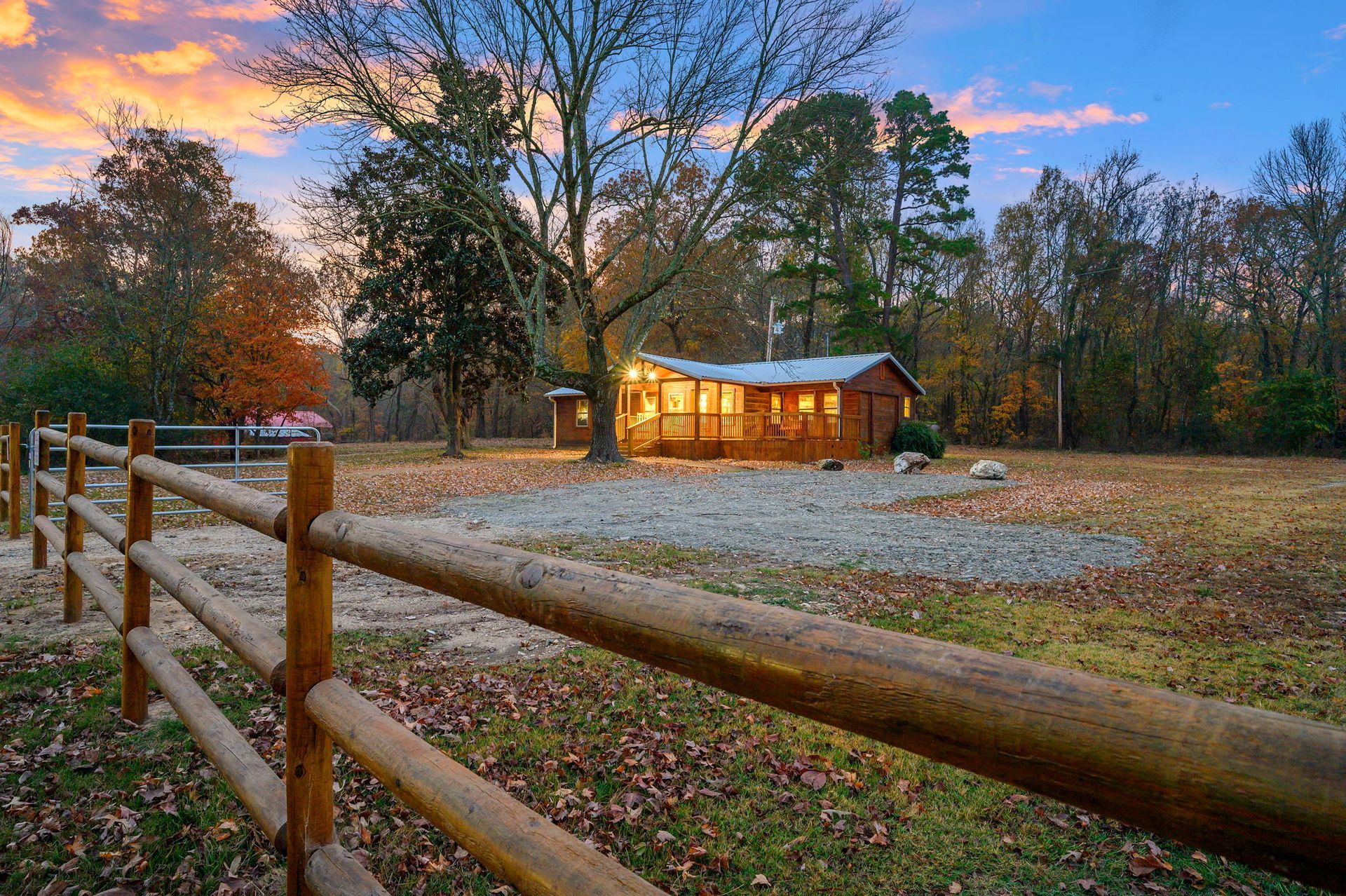 A rustic wooden cabin illuminated at sunset, seen beyond a wooden fence in a yard scattered with autumn leaves.