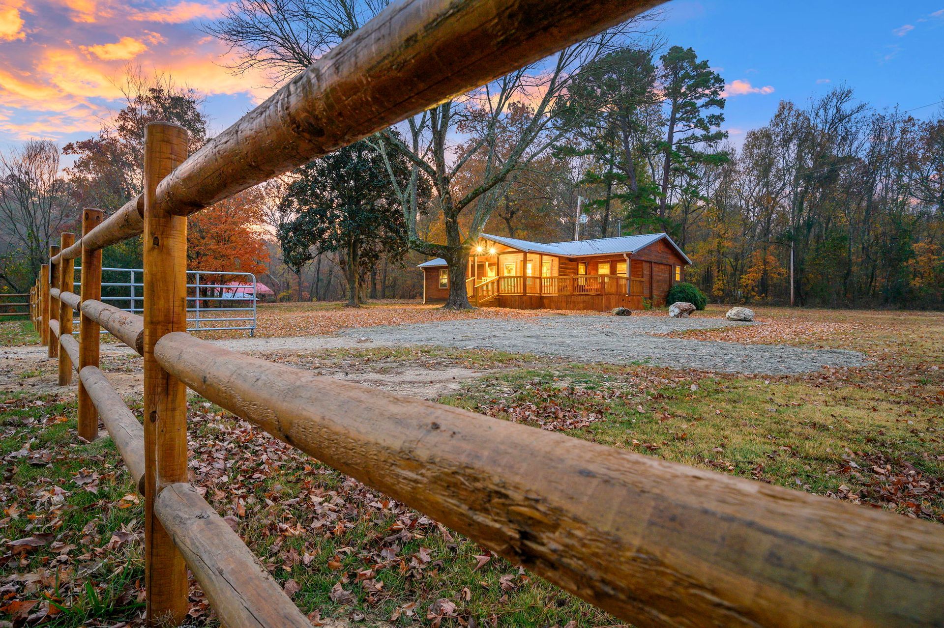 A rustic wooden cabin with illuminated windows sits at sunset behind a split-rail fence in a leaf-covered field.