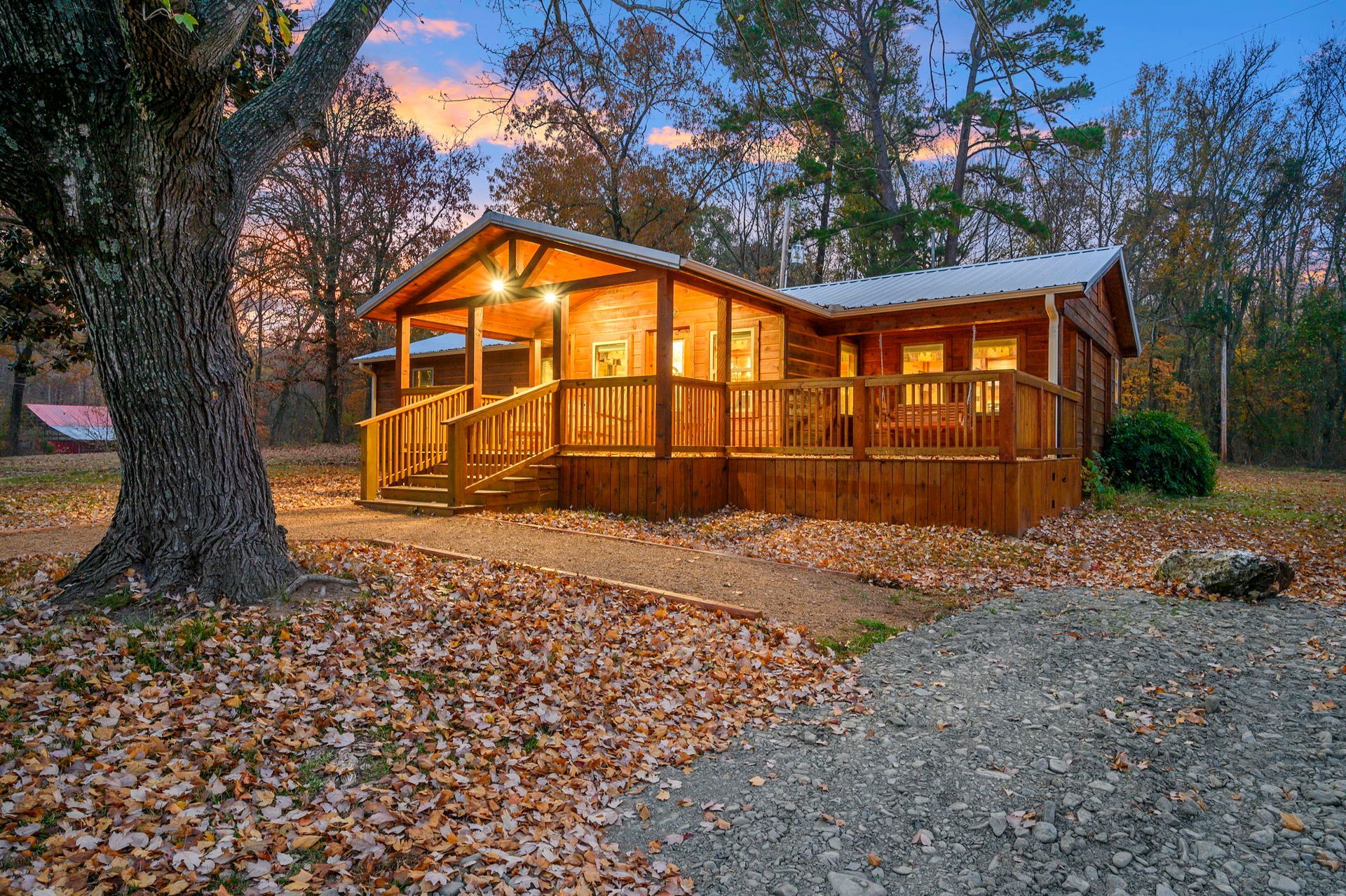 A rustic wooden cabin with a covered deck and front porch, surrounded by fall foliage under a sunset sky.