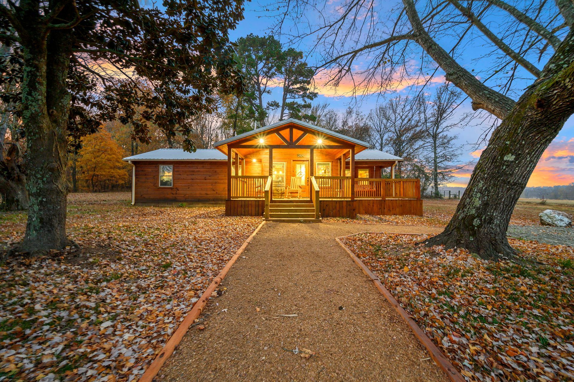 A wooden house with a covered porch at sunset, approached by a gravel path surrounded by autumn leaves and trees.
