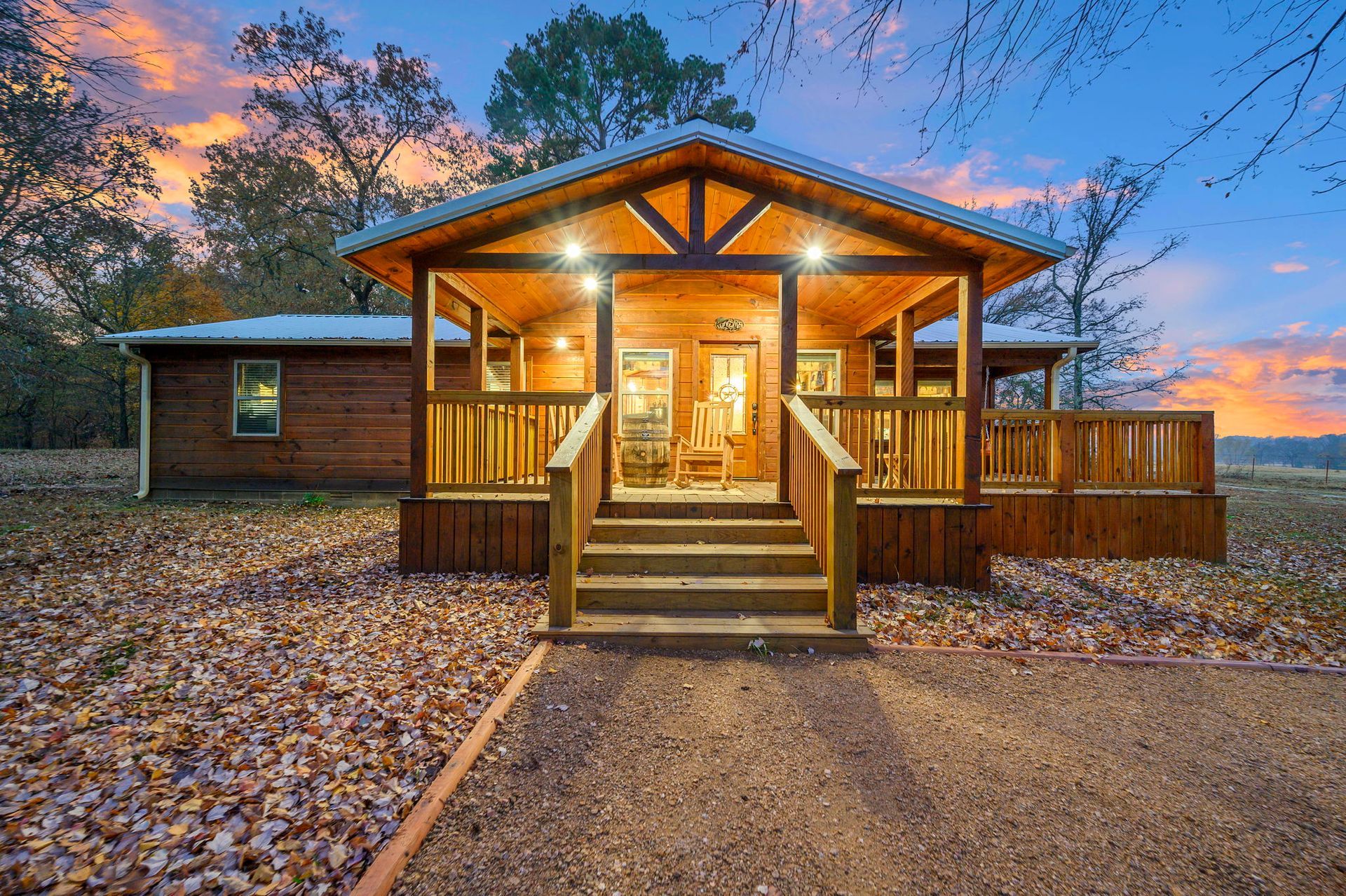 A wooden cabin with a covered front porch and stairs at dusk, set against a backdrop of trees and a sunset sky.