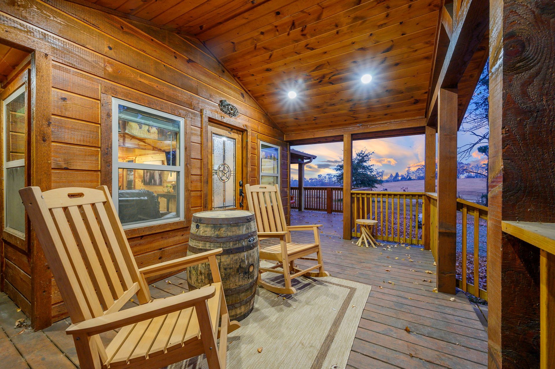 Two wooden rocking chairs and a barrel side table sit on a porch covered by a wooden roof at dusk.