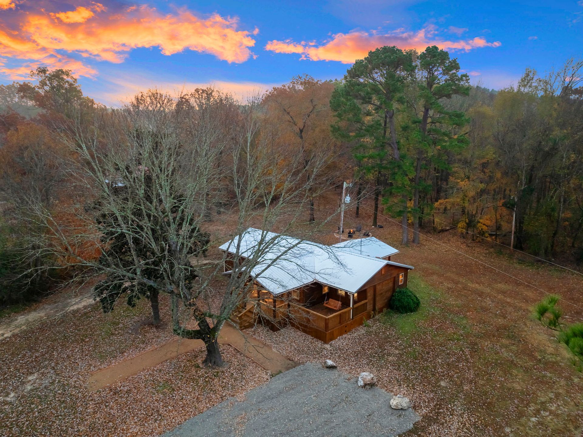 An elevated view of a wooden cabin with a silver metal roof nestled in a wooded area under a sunset sky.