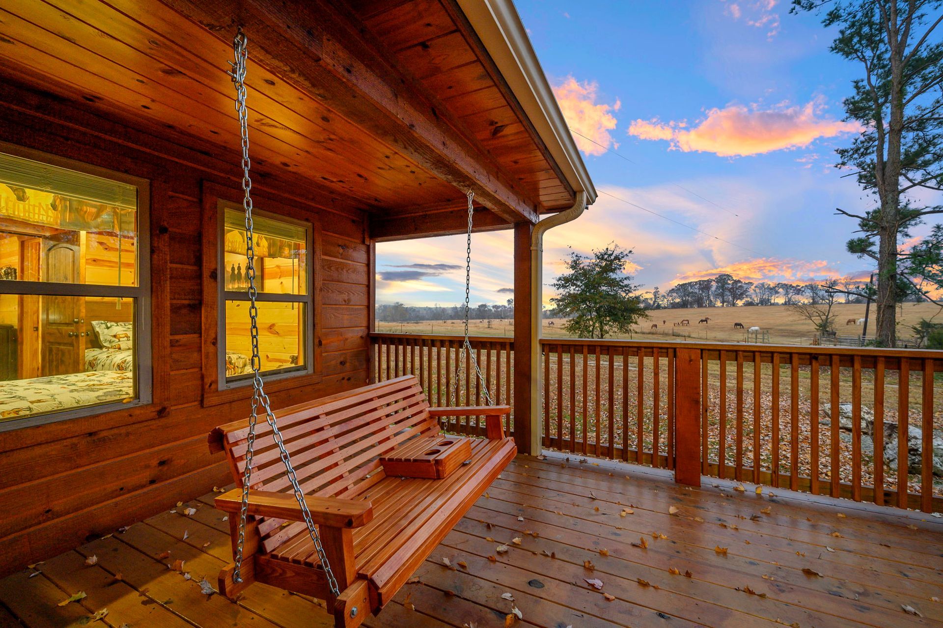 A wooden porch swing hangs on the deck of a rustic log cabin, overlooking a field at sunset.