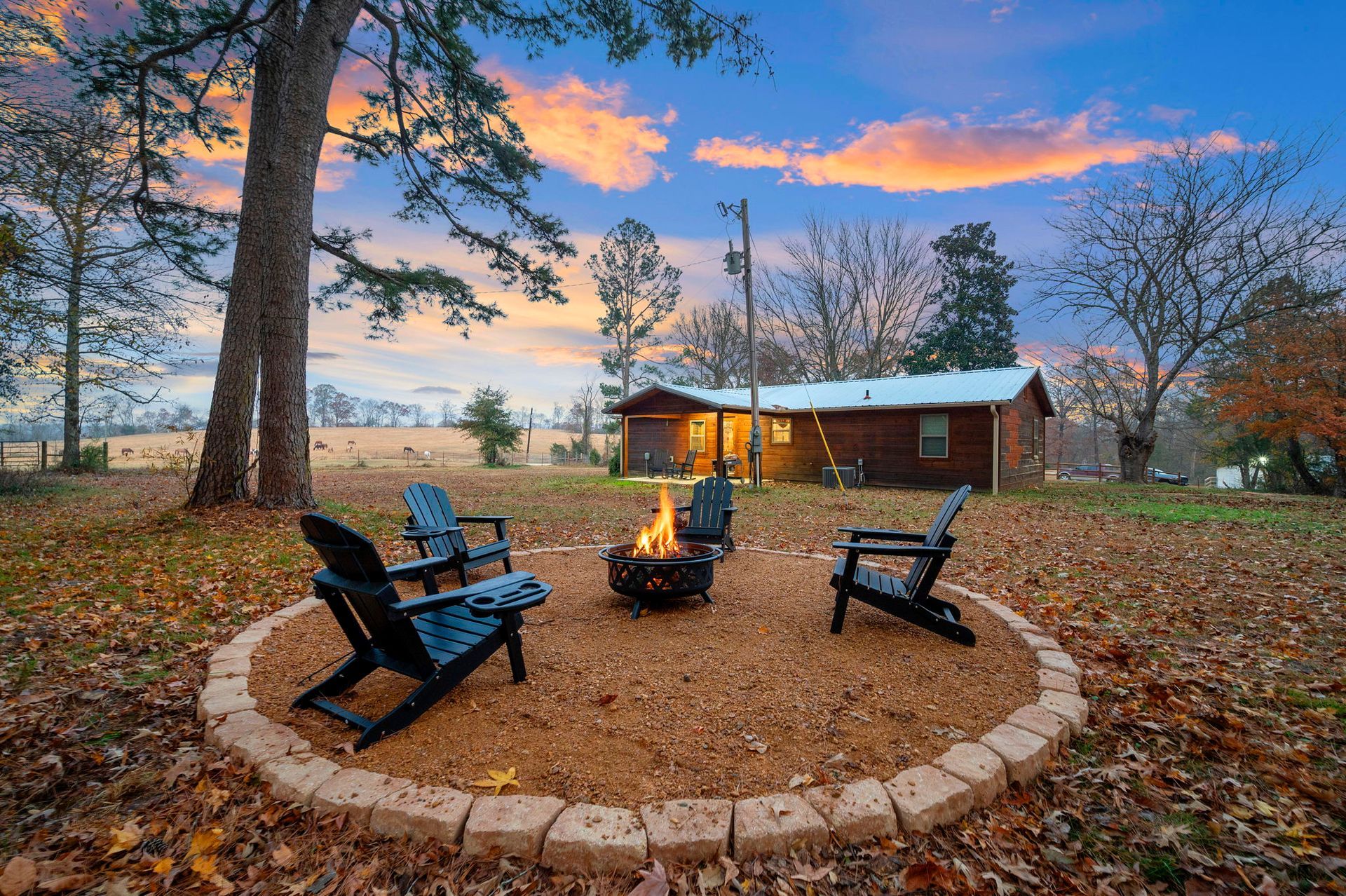 A fire pit with four black Adirondack chairs sits on a circular stone patio at dusk in front of a rustic wooden cabin.