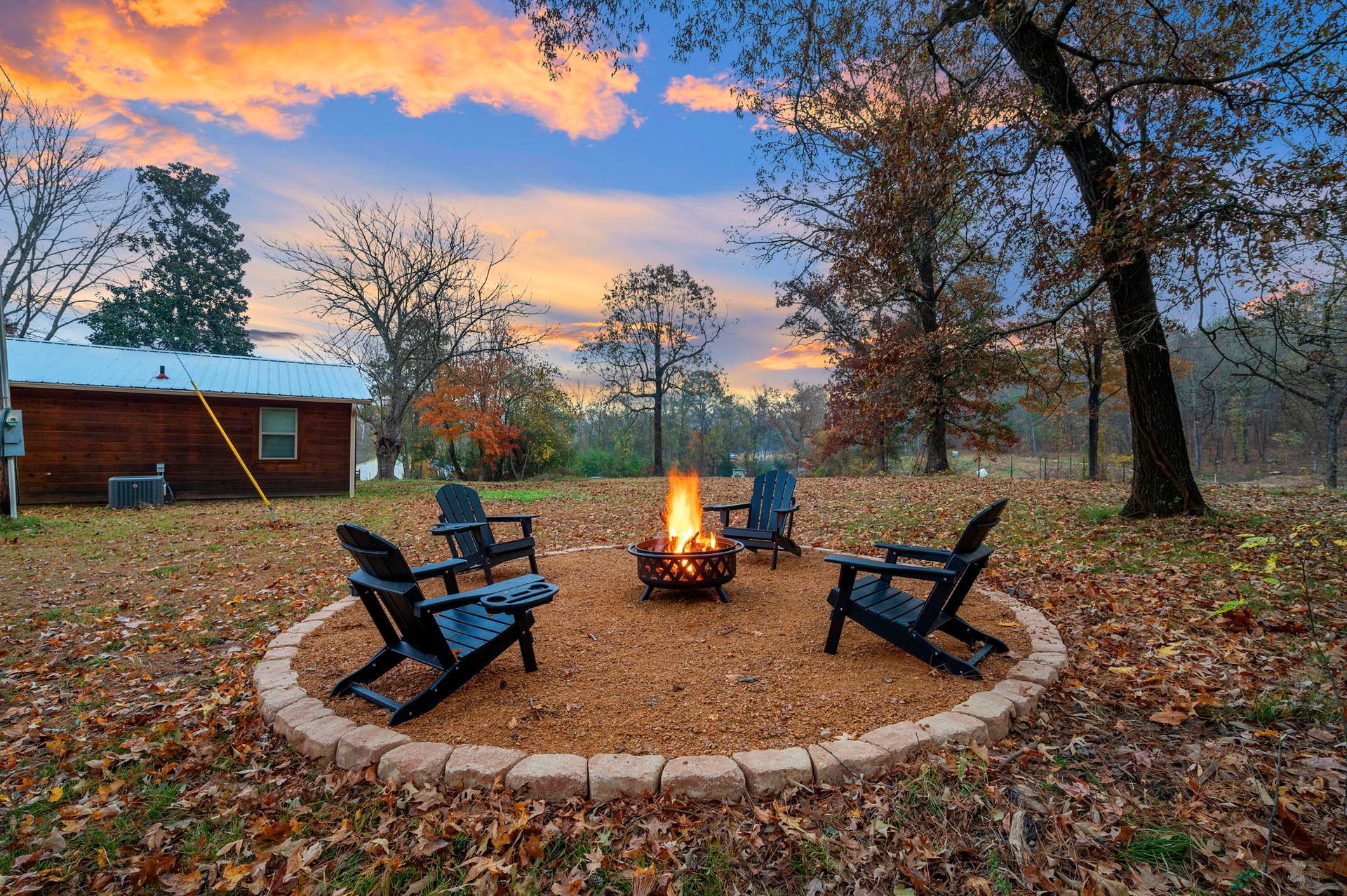 A circular fire pit surrounded by four dark chairs on a wood-mulched area in a yard filled with autumn leaves at sunset.